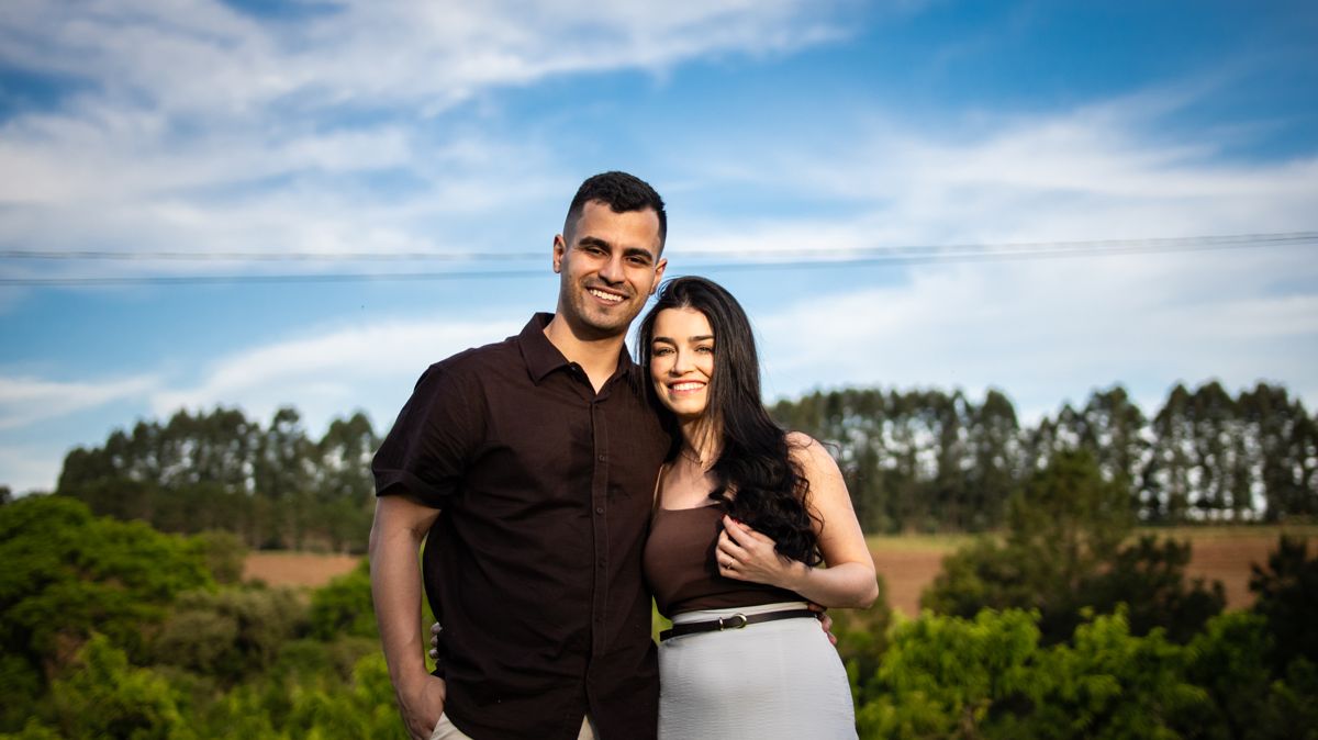 Juliana e Matheus abraçados, sorrindo, sob o céu azul